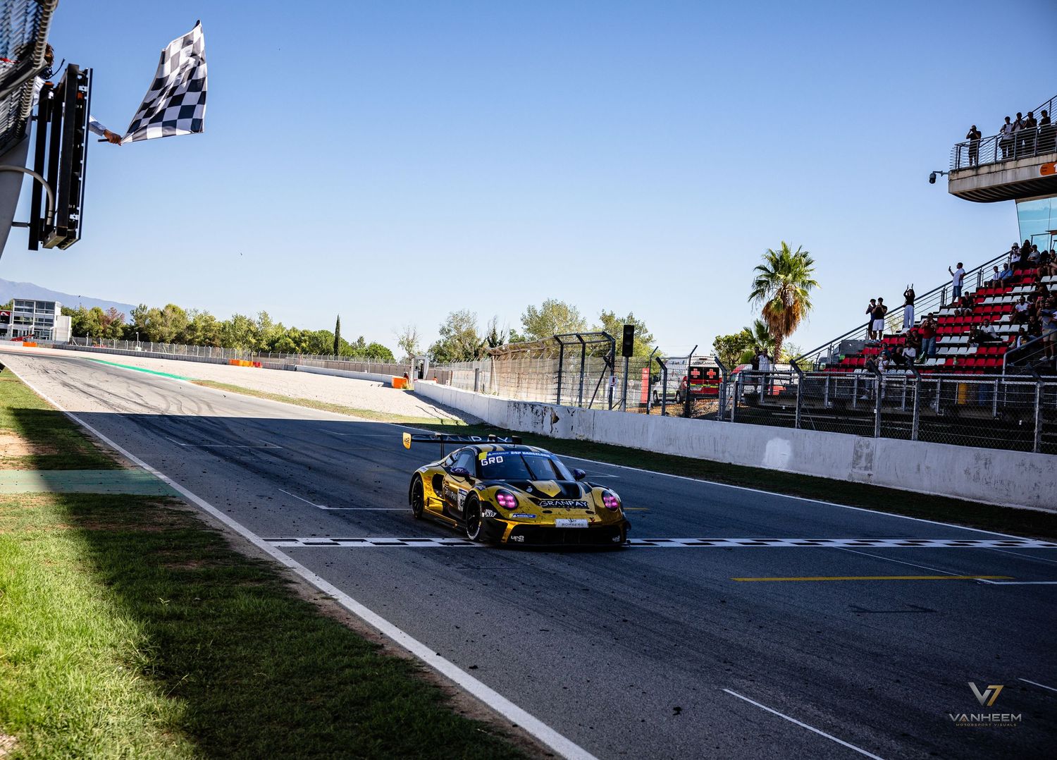 Yellow racing car crosses the finish line under checkered flag at a sunny motorsport circuit.
