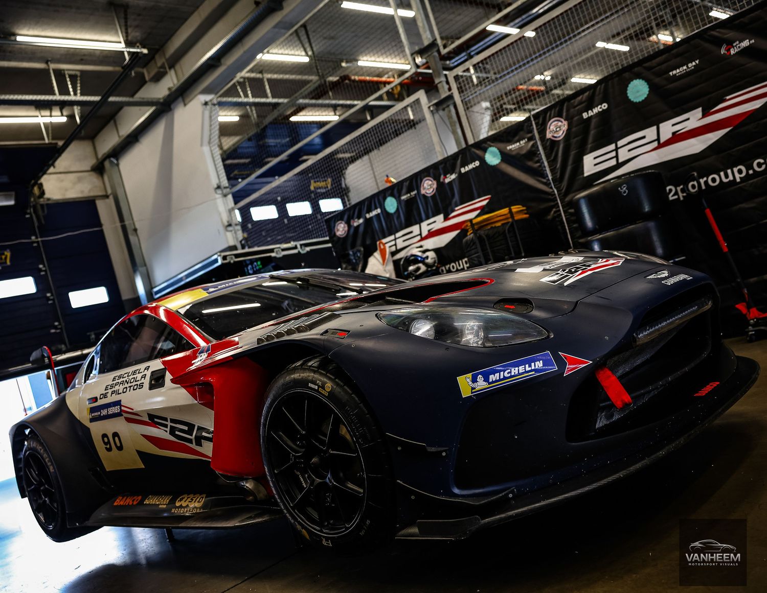 A sleek racing car with red, white, and blue livery parked in a dimly lit pit garage at a motorsport event.