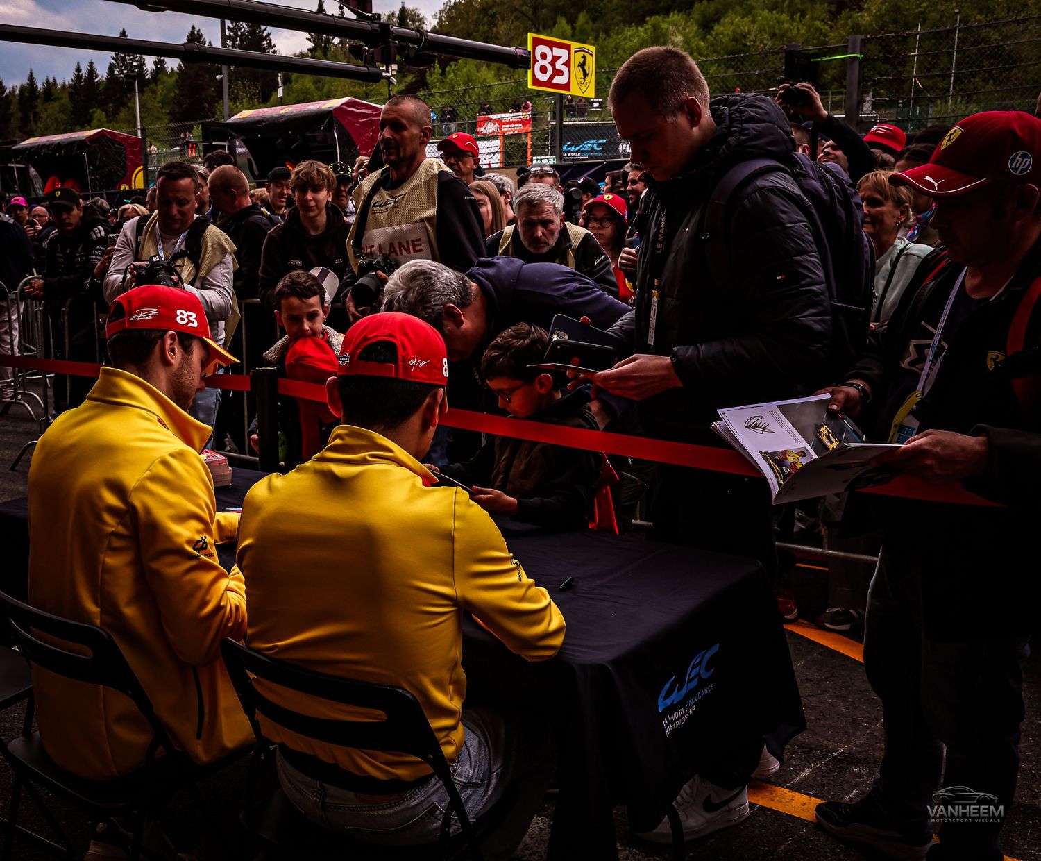 Racing team members in yellow jackets sign autographs for fans wearing red caps at a motorsport event.