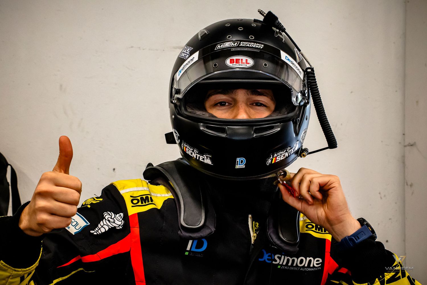Racing driver in Bell helmet and black suit gives thumbs up, wearing sponsor logos including JeSimone.
