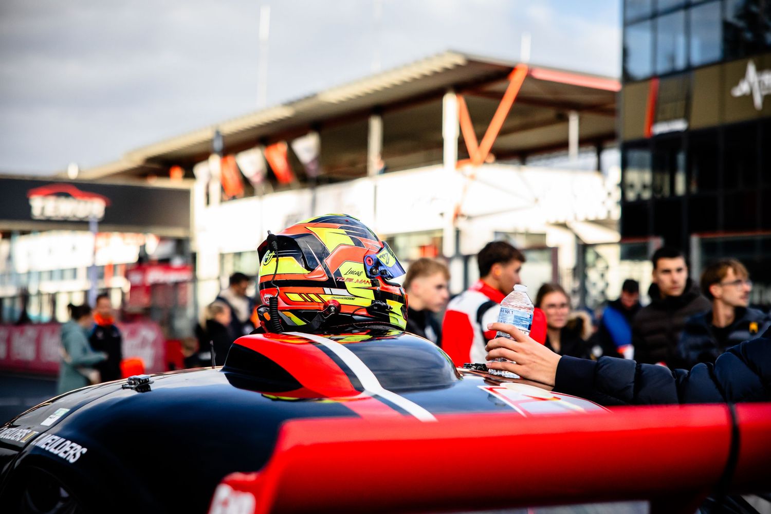 A racing driver in a red and yellow helmet sits in a Formula car at a motorsport circuit paddock.