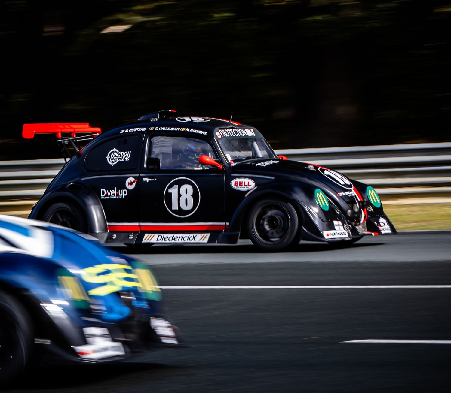 Black VW Beetle race car number 18 competing on a racing circuit alongside a blue car, captured with motion blur effect.