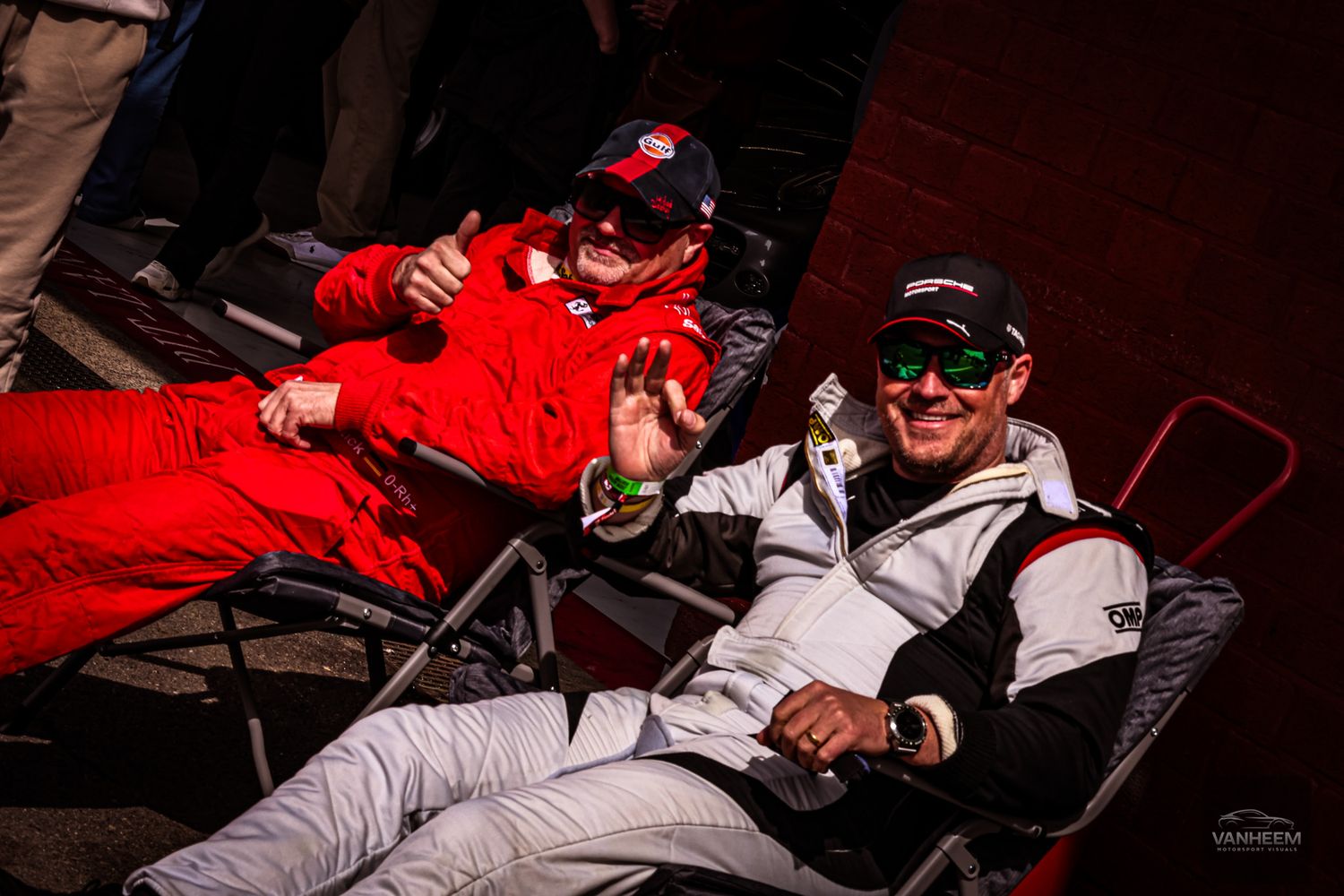 Two racing drivers in motorsport gear relax in folding chairs, smiling and giving thumbs up at a race event.