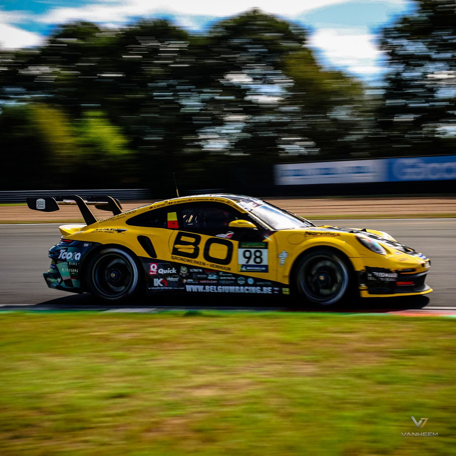 Yellow and black Porsche racing car number 98 speeding on a motorsport circuit track with motion blur background.