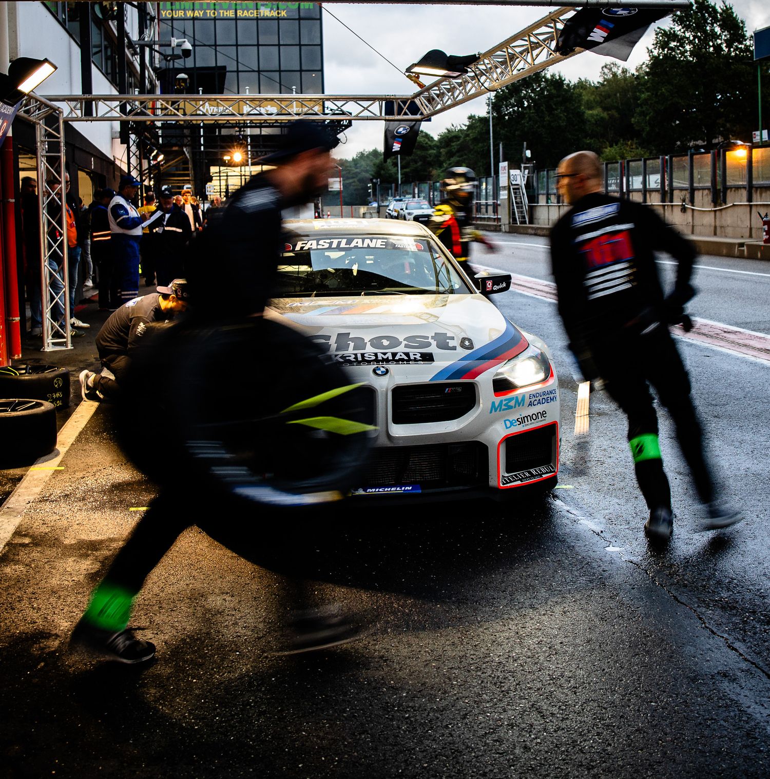 BMW race car in pit lane with crew members rushing during a wet motorsport event at night.