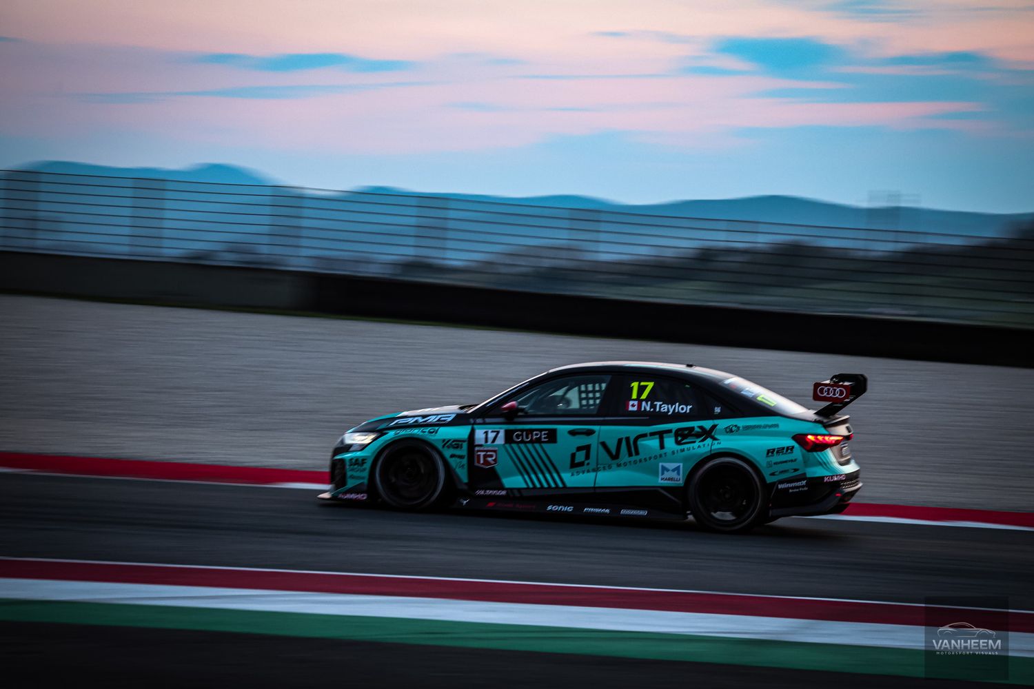 A row of race cars speeding along a track during sunset with dramatic sky and mountains in the background.