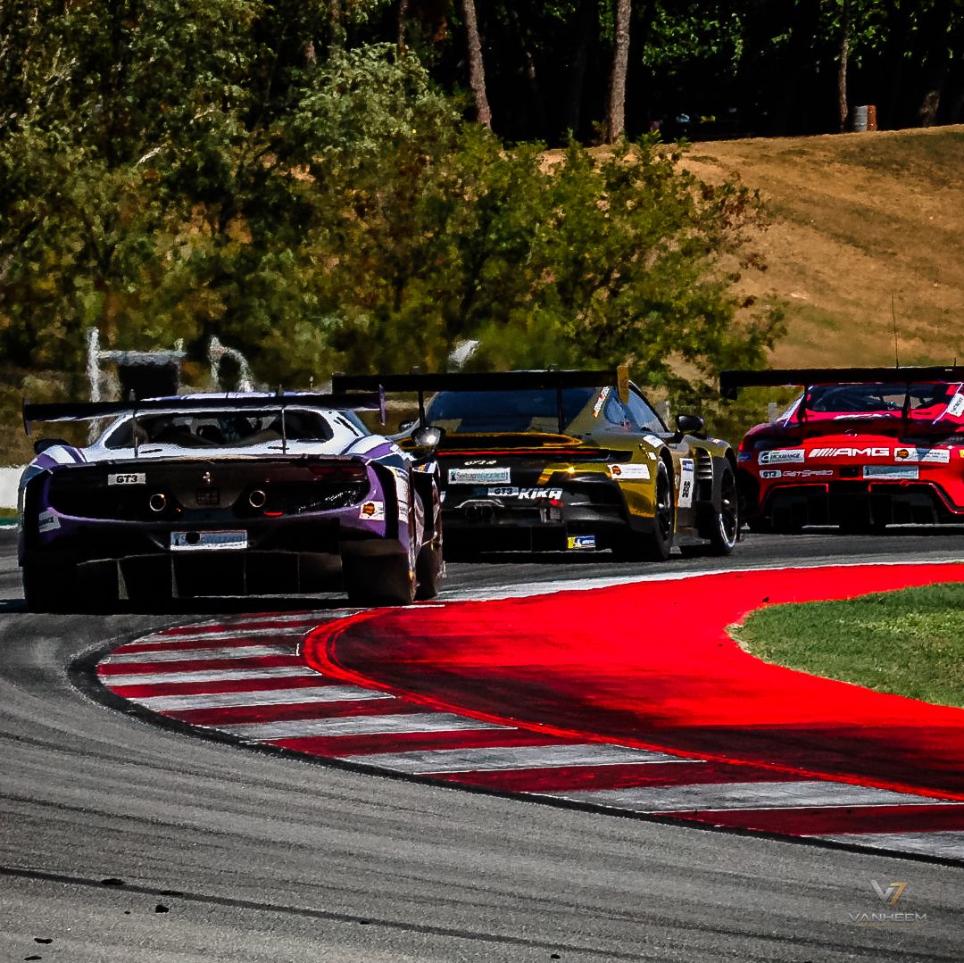 Three GT race cars navigate a red curb corner on a scenic motorsport track surrounded by lush green trees.