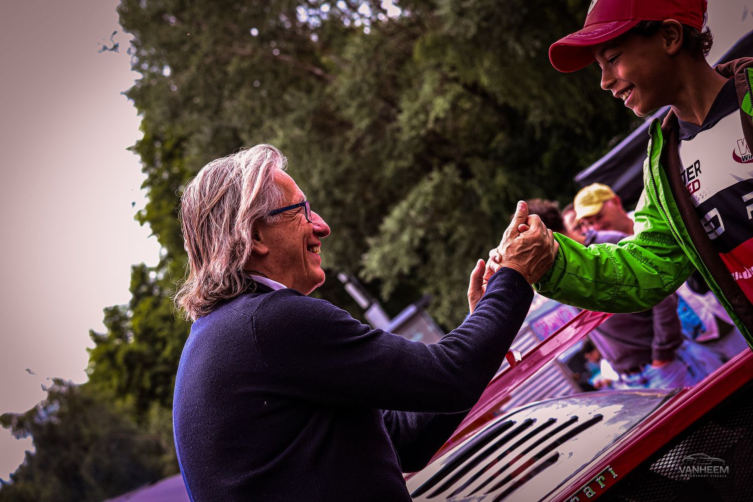 Two people shaking hands at an outdoor racing event, one wearing a green jacket and red cap.