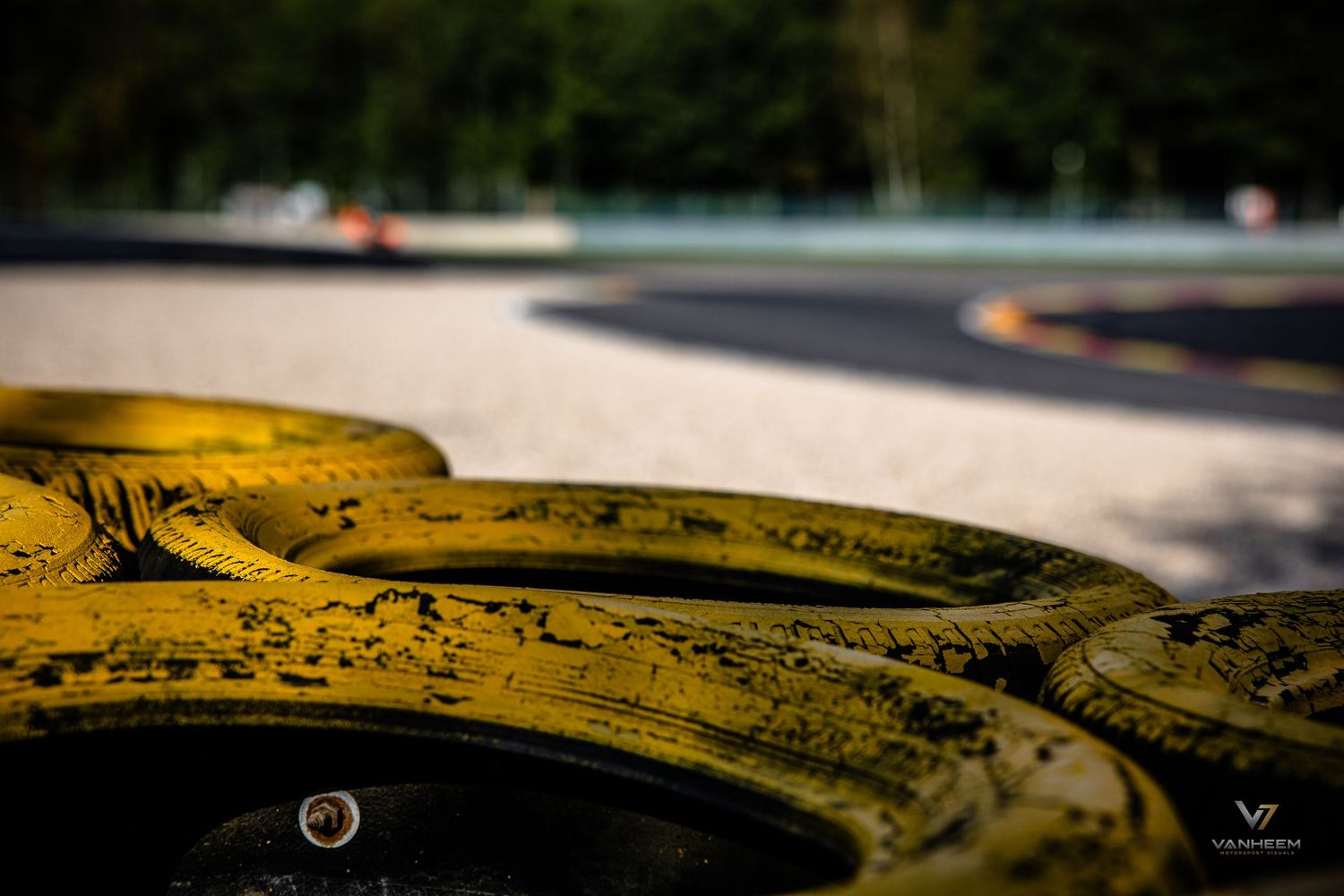 Yellow-painted tires line a racing circuit track barrier, with a blurred go-kart track visible in the background.