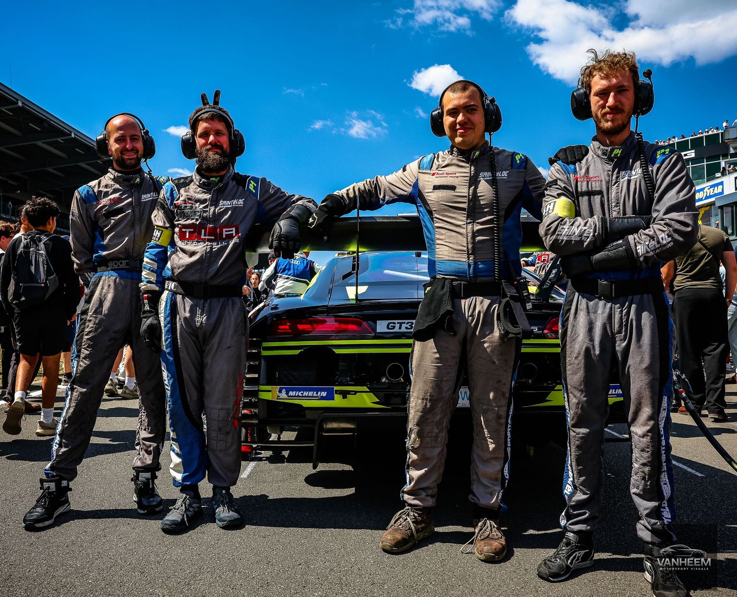 Four racing drivers in team uniforms pose together on a motorsport grid beside a race car on a sunny day.