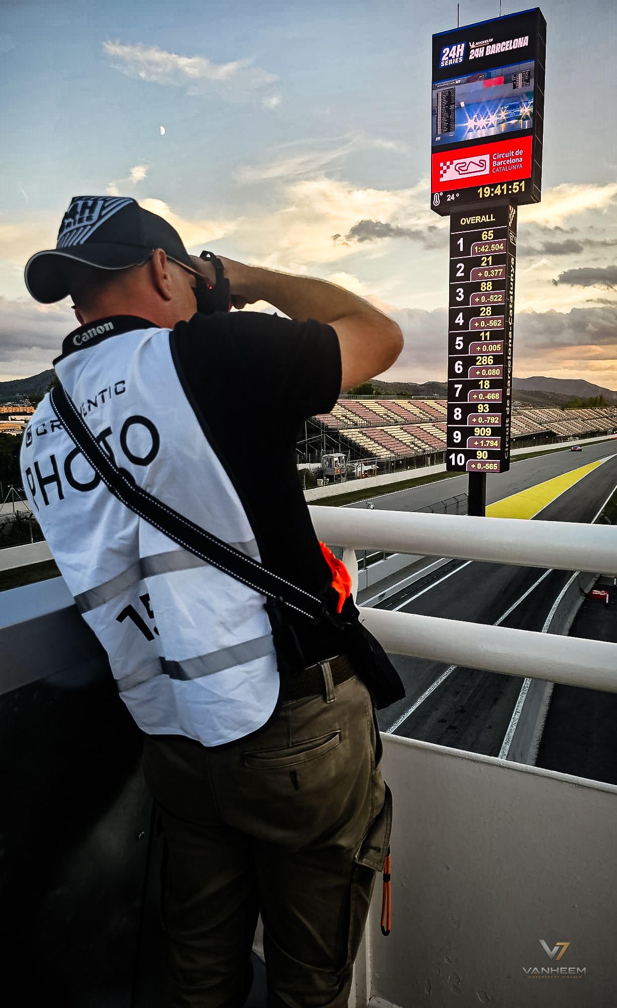 Photographer with Canon camera and PHOTO vest shoots at a racing circuit with a timing board at sunset.