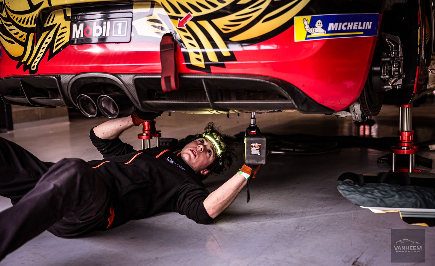 A mechanic lying under a red race car with Mobil1 and Michelin sponsorship logos, performing maintenance in a garage pit lane.