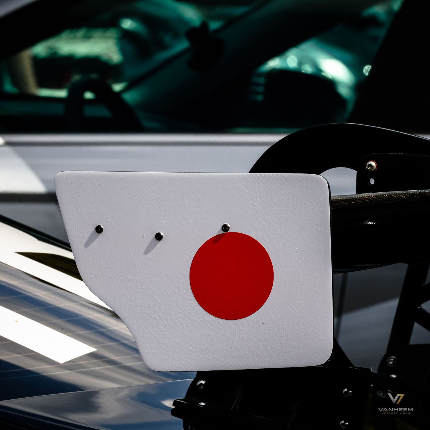 Close-up of a Japanese flag decal on a white car spoiler wing, mounted on a vehicle hood outdoors.