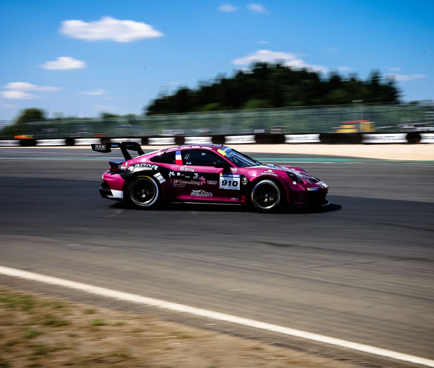 Pink racing car number 910 speeding on a sunny race track with trees in the background.