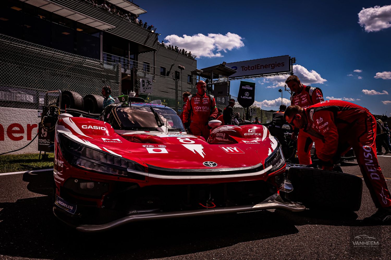 Red Toyota GR racing car in pit lane with crew members performing tire change at sunny motorsport event.