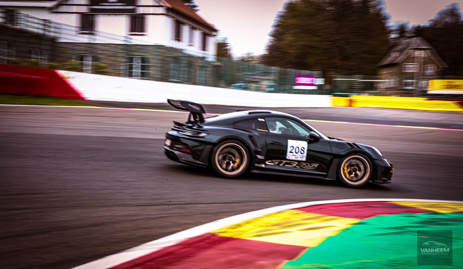 Black Porsche GT3 race car with gold wheels speeding on Spa-Francorchamps circuit track during a motorsport event.