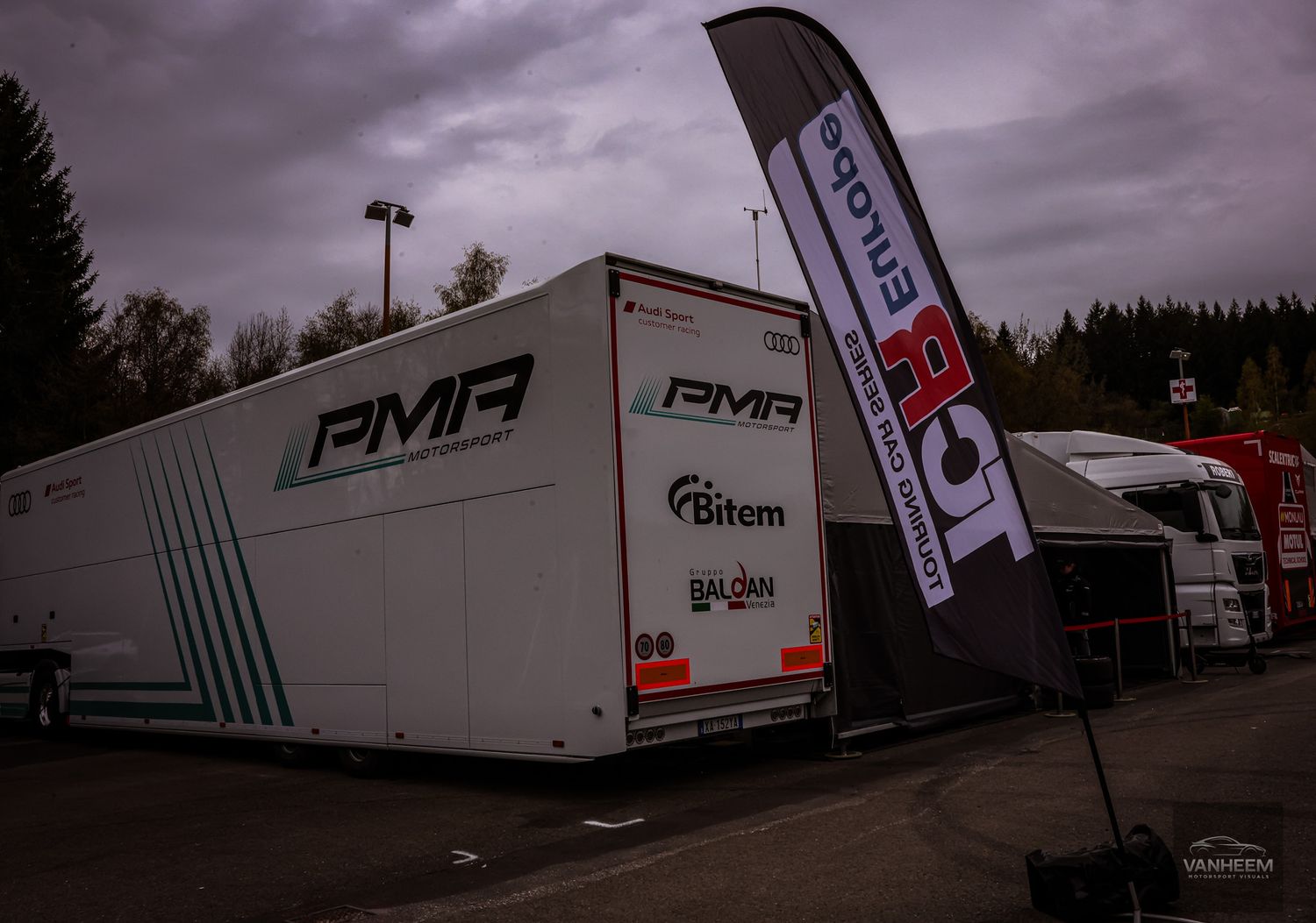PMA racing team trailer and branded banner displayed at a motorsport event paddock area on an overcast day.