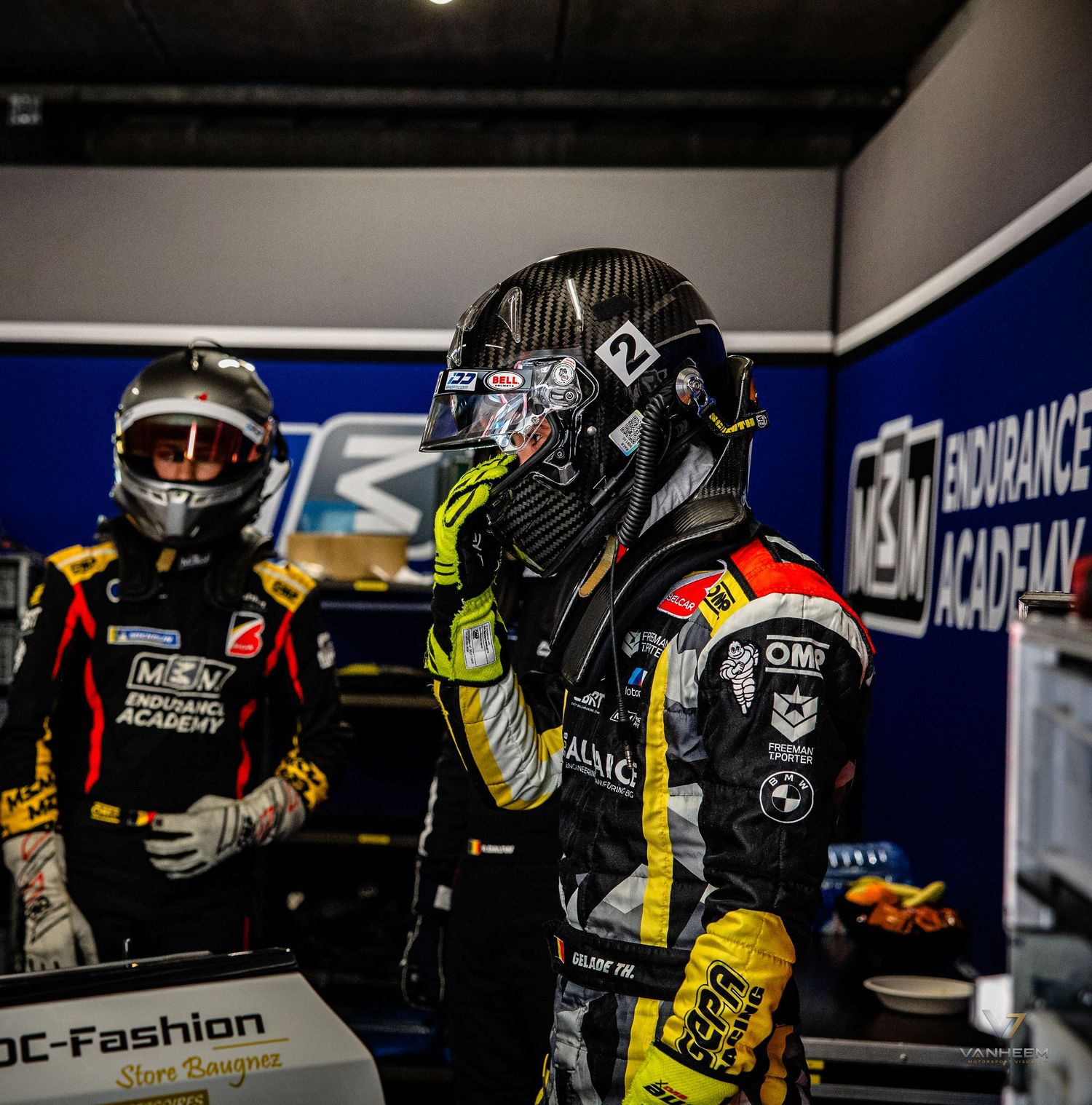 Two racing drivers in helmets and suits stand in a pit garage at an endurance racing academy event.