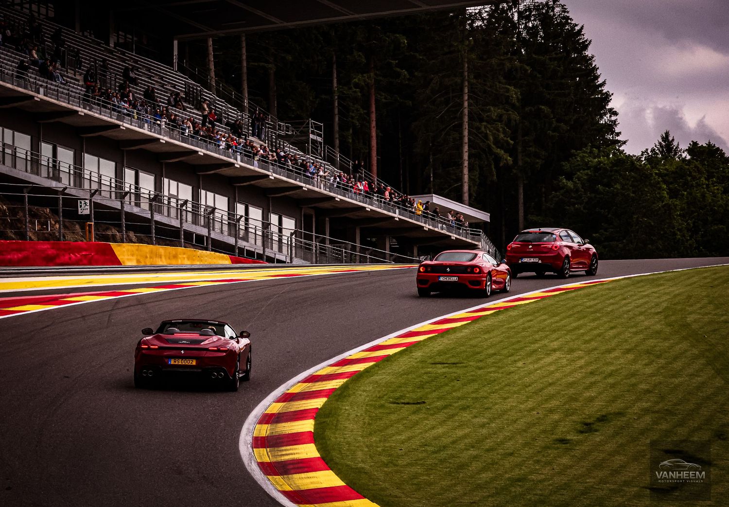 Red Ferrari sports cars racing on a wet circuit track with grandstands and lush green trees in background.