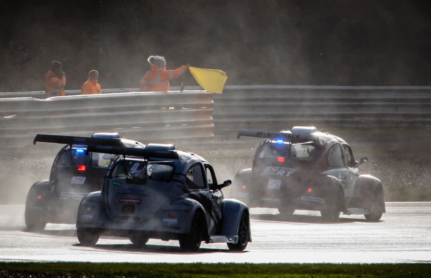 Vintage race cars compete on a wet track in rainy conditions, with marshals waving yellow flags in the background.