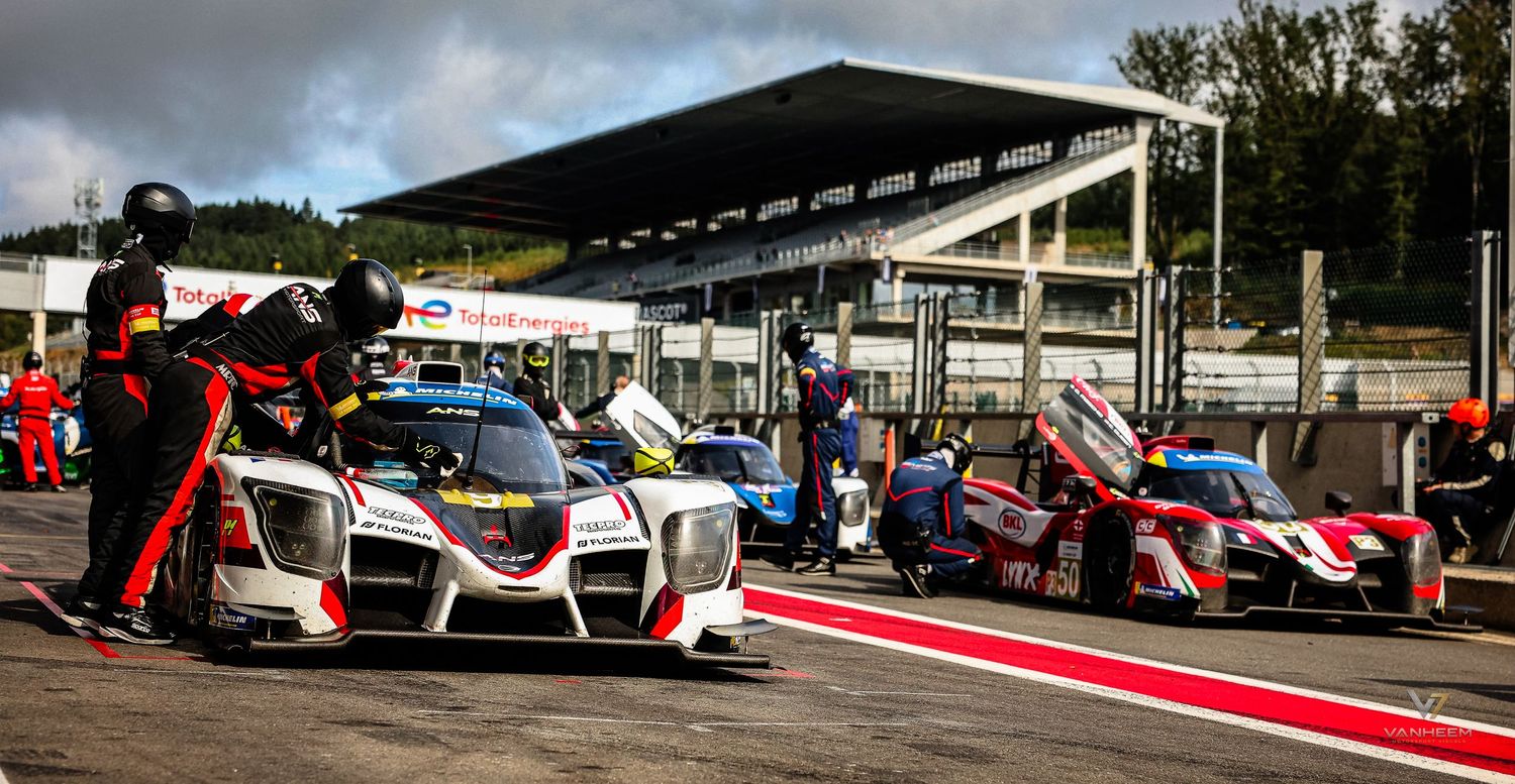 Racing cars in pit lane at a motorsport circuit during a cloudy day, with crew members in red suits attending vehicles.