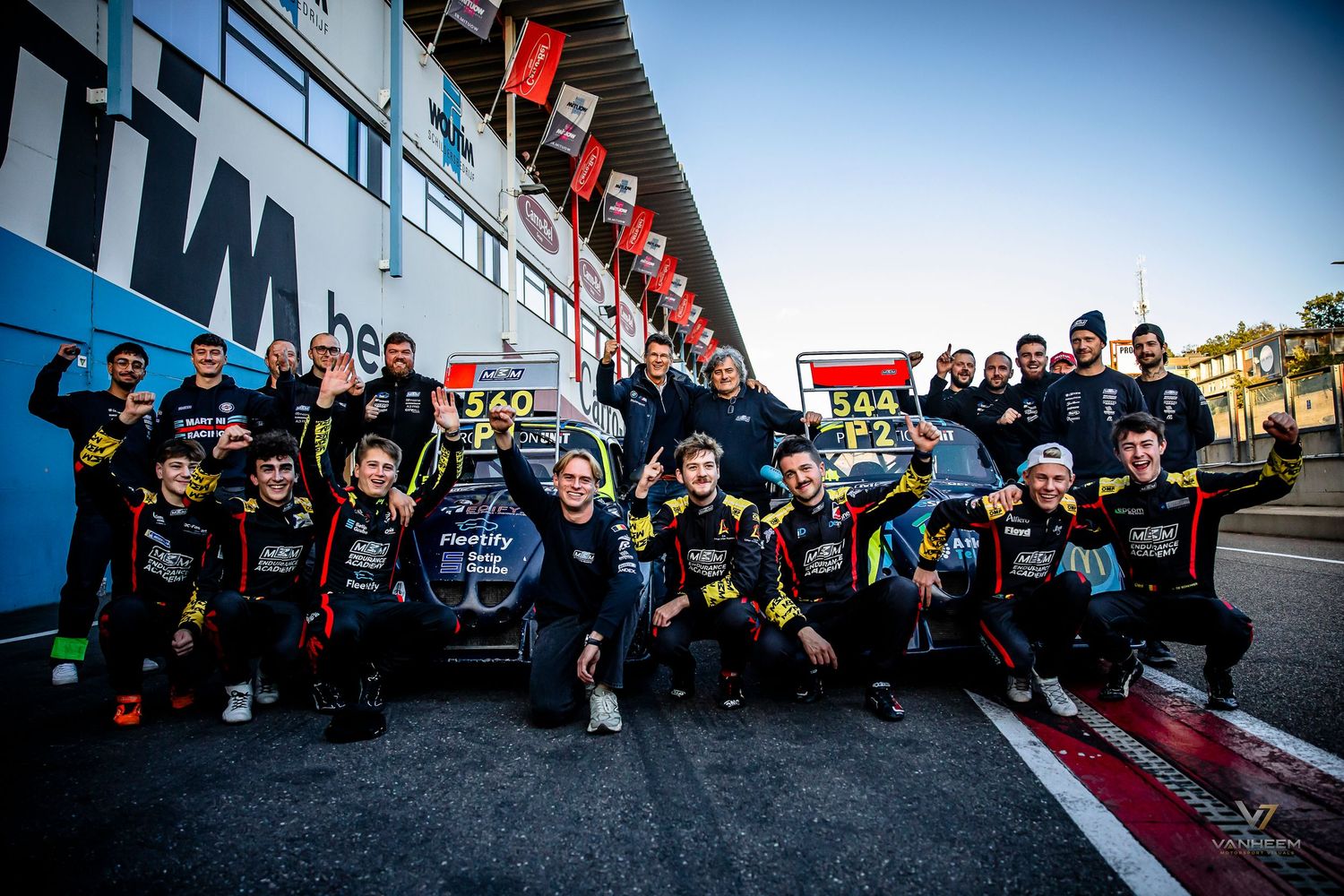 Racing team celebrates victory together at pit lane, wearing matching black and yellow uniforms at a motorsport circuit.
