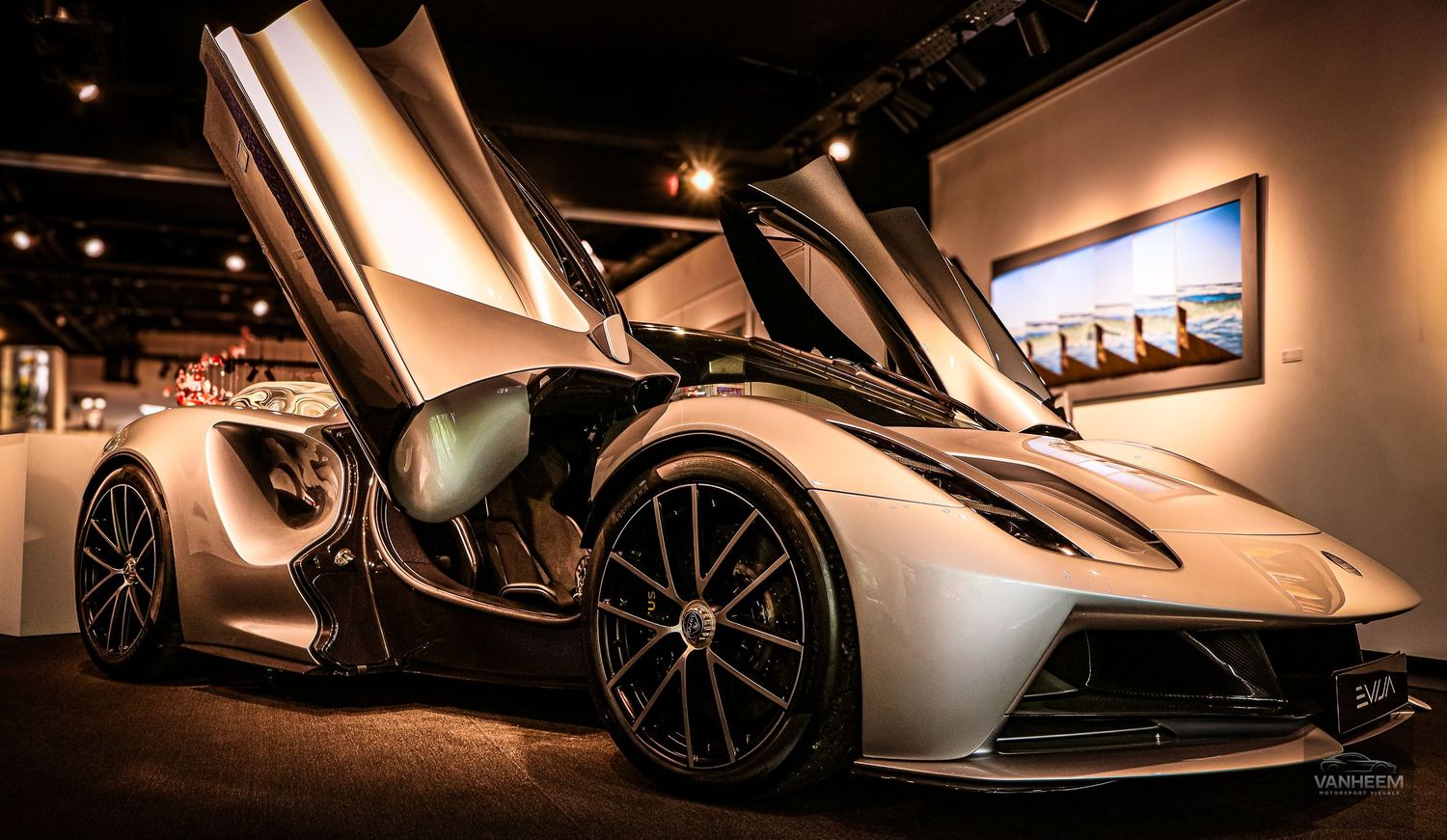 Silver supercar with scissor doors open on display in a showroom, showcasing its sleek aerodynamic design.