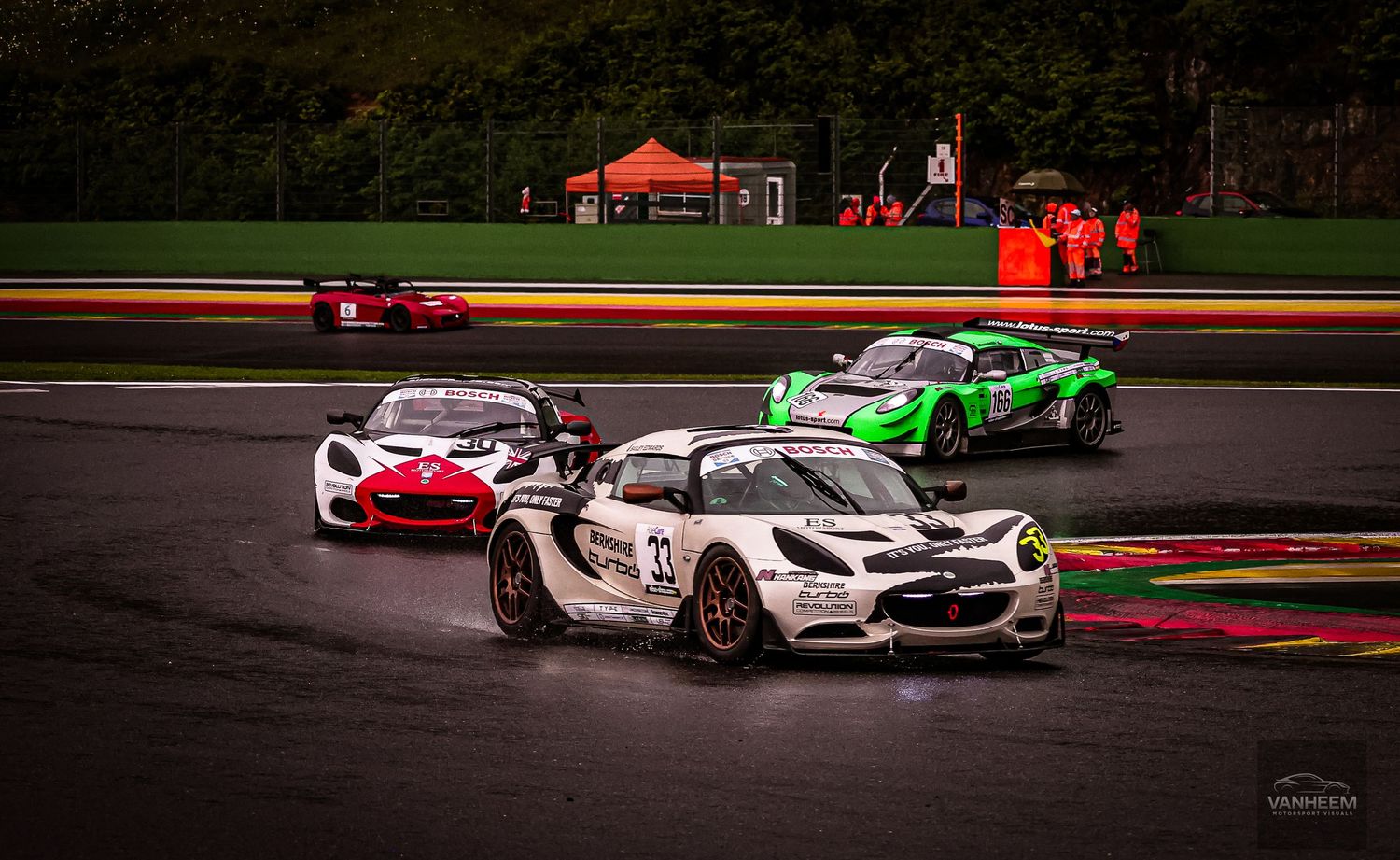 Racing cars compete on a wet track at Spa-Francorchamps, with a Lotus Elise leading the pack in rainy conditions.