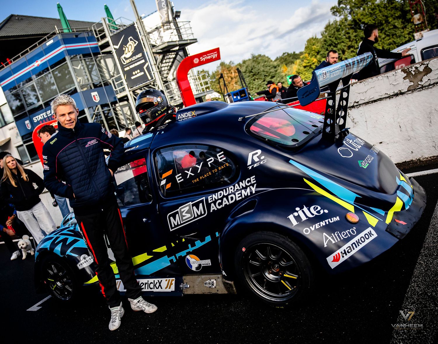 Racing driver poses beside a dark blue GT race car at a motorsport endurance academy event on a city street circuit.
