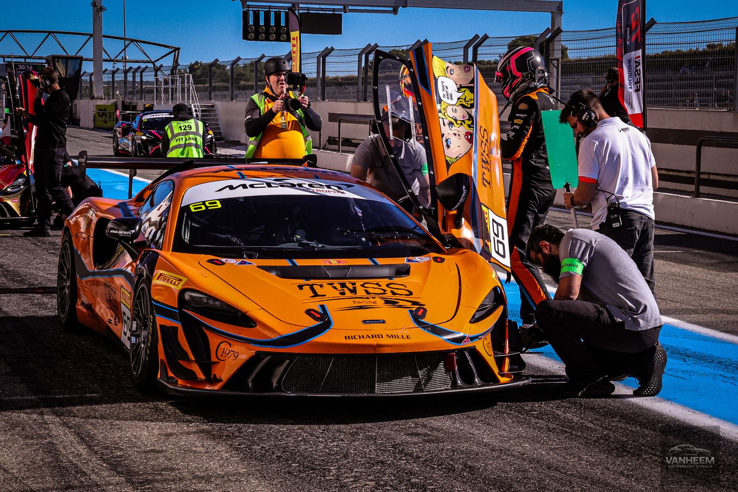 Orange McLaren GT3 race car being serviced by pit crew during race day.