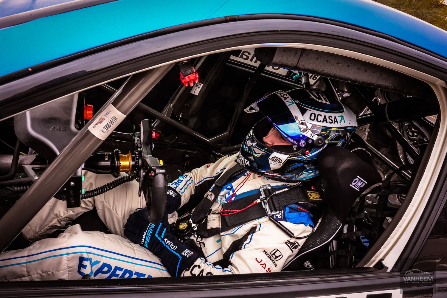 Racing driver seated in a blue race car cockpit wearing helmet and safety gear with sponsor logos visible.