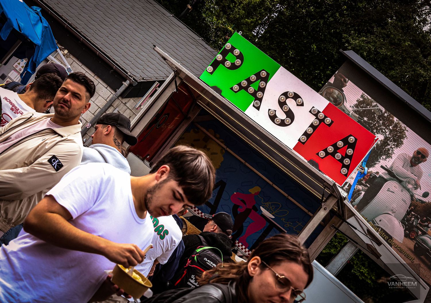 Crowd of people at an outdoor Italian pasta food stall with a colorful green, white, and red sign.