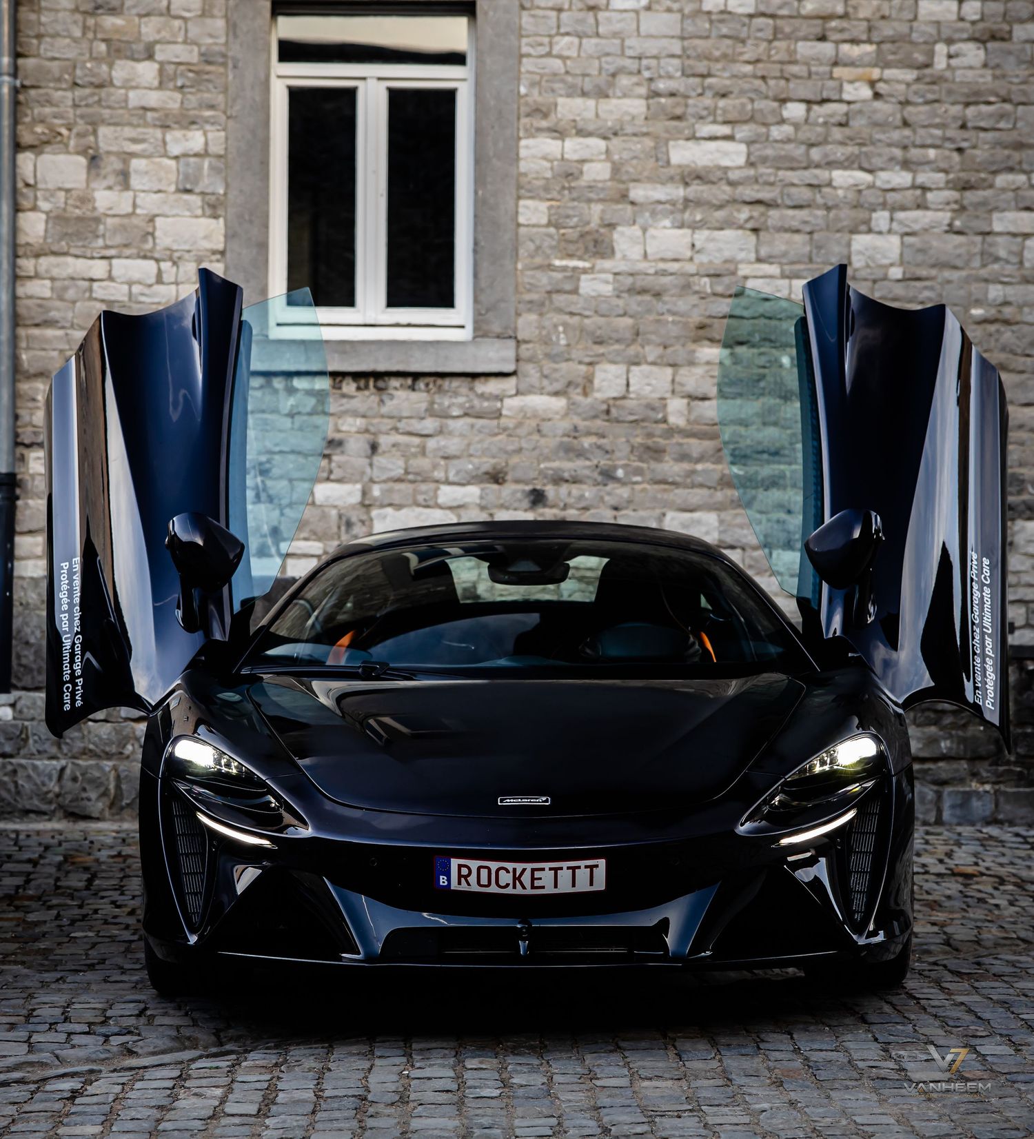 Black McLaren supercar with dihedral doors open, license plate ROCKETTT, parked on cobblestones before a stone building.