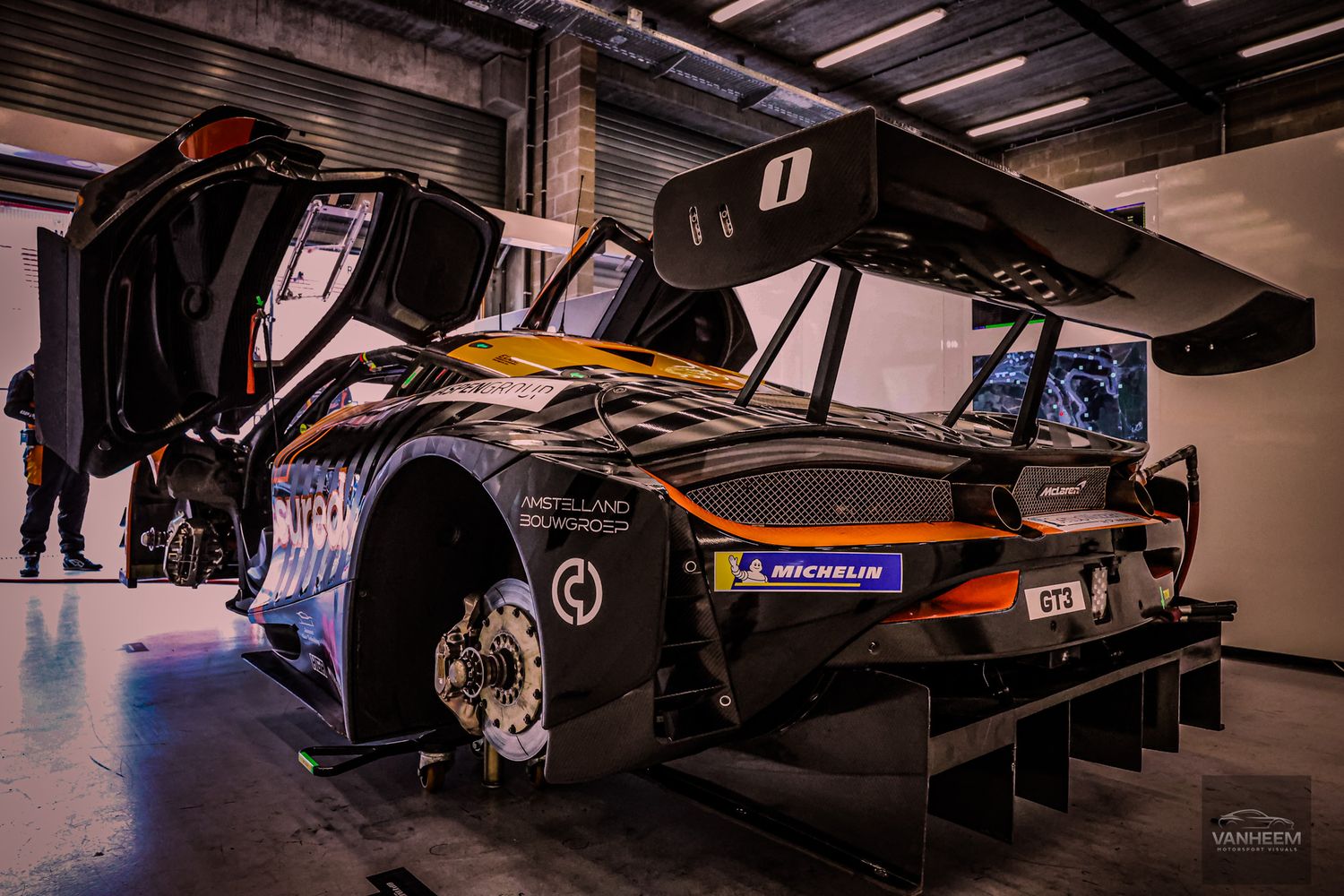 A McLaren GT race car with Michelin tires sits in a garage, featuring a large rear wing and racing livery.