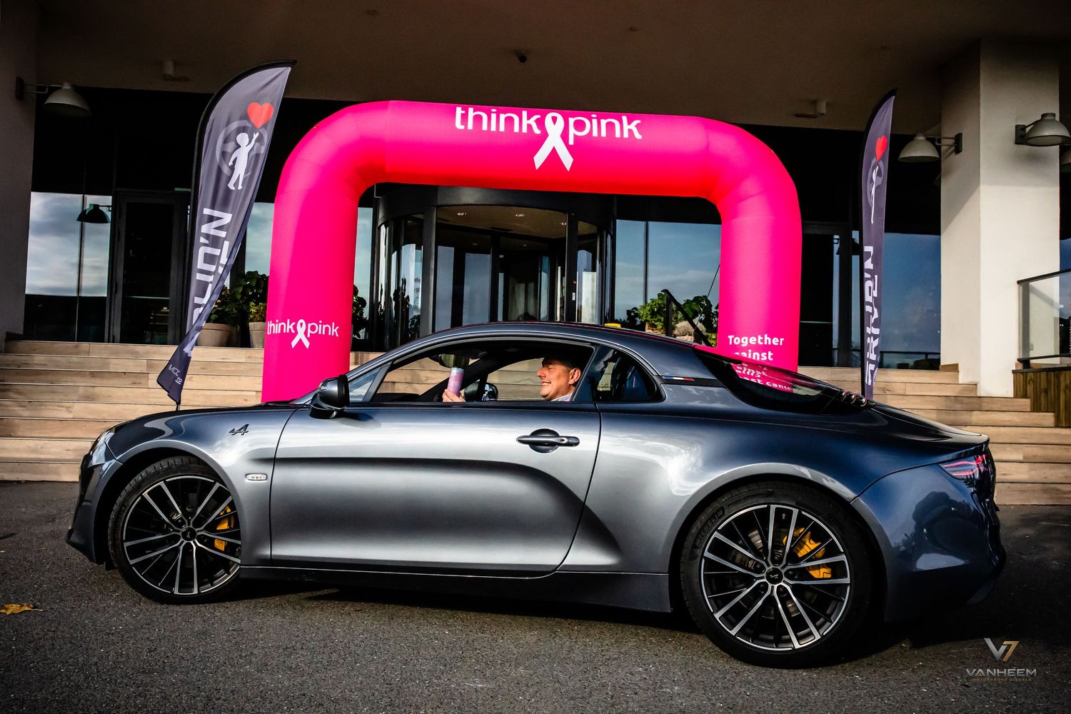 Gray sports car parked in front of a pink Think Pink breast cancer awareness arch at an outdoor event.