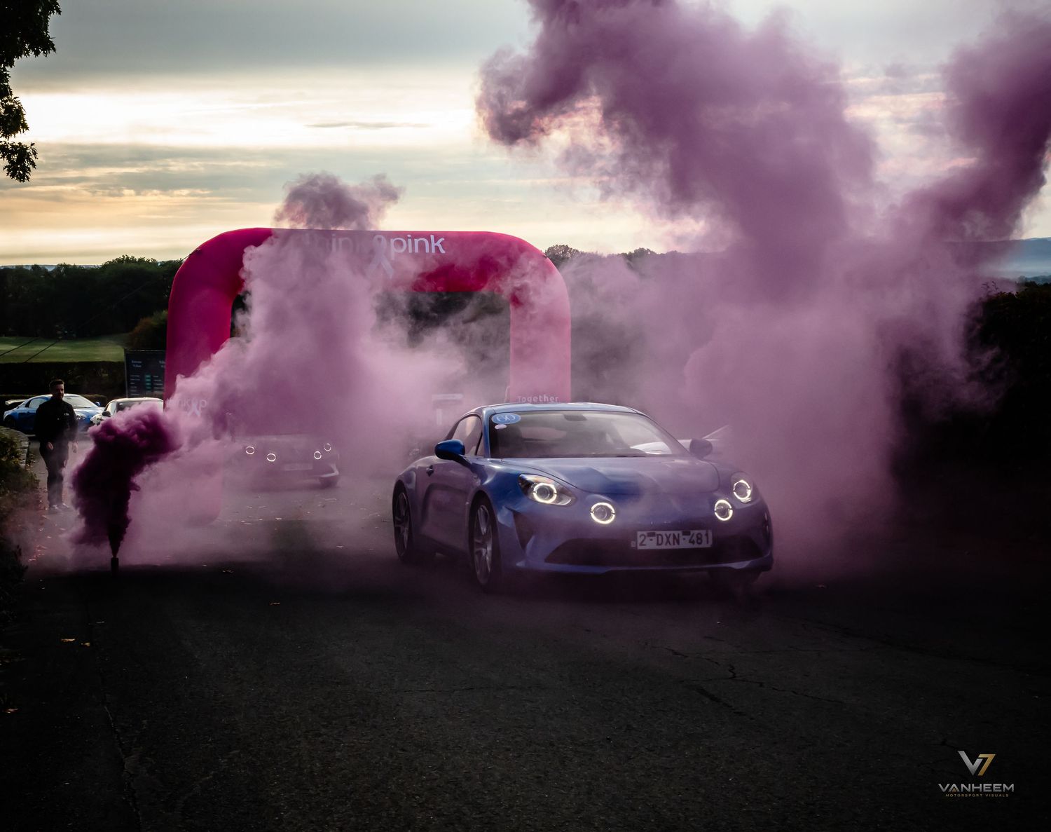 A sleek blue sports car emerges dramatically through vibrant pink smoke at an outdoor automotive event at dusk.