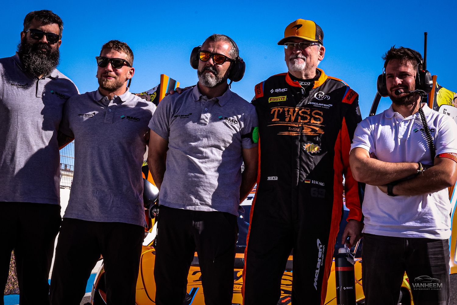 Racing team members gather at a motorsport event wearing team uniforms and gear.