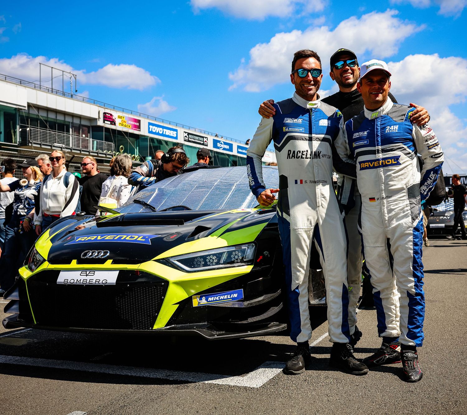 Three racing drivers pose beside an Audi GT3 race car at a motorsport circuit on a sunny day.