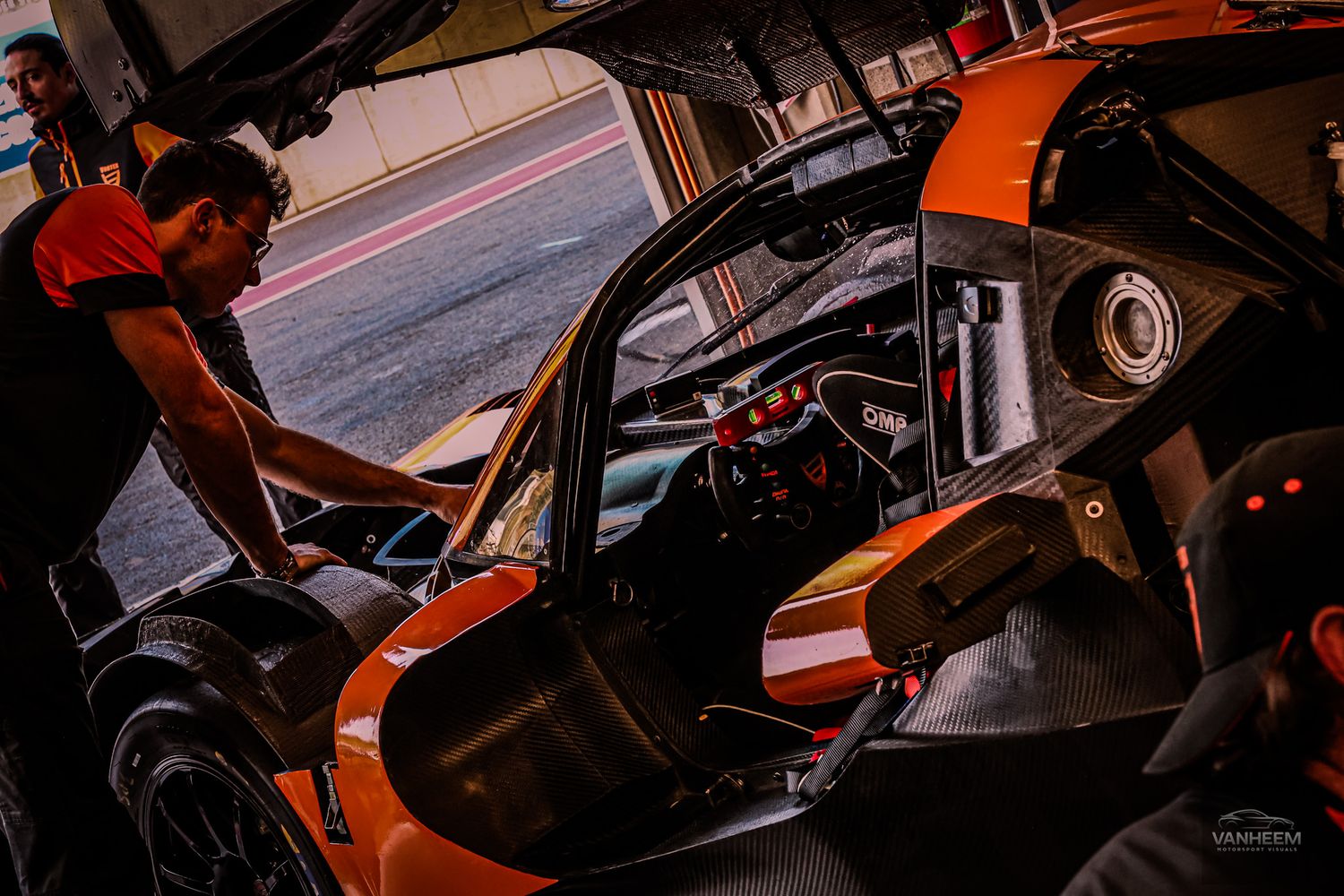 Mechanic working on an orange racing car in a pit garage during a motorsport event.