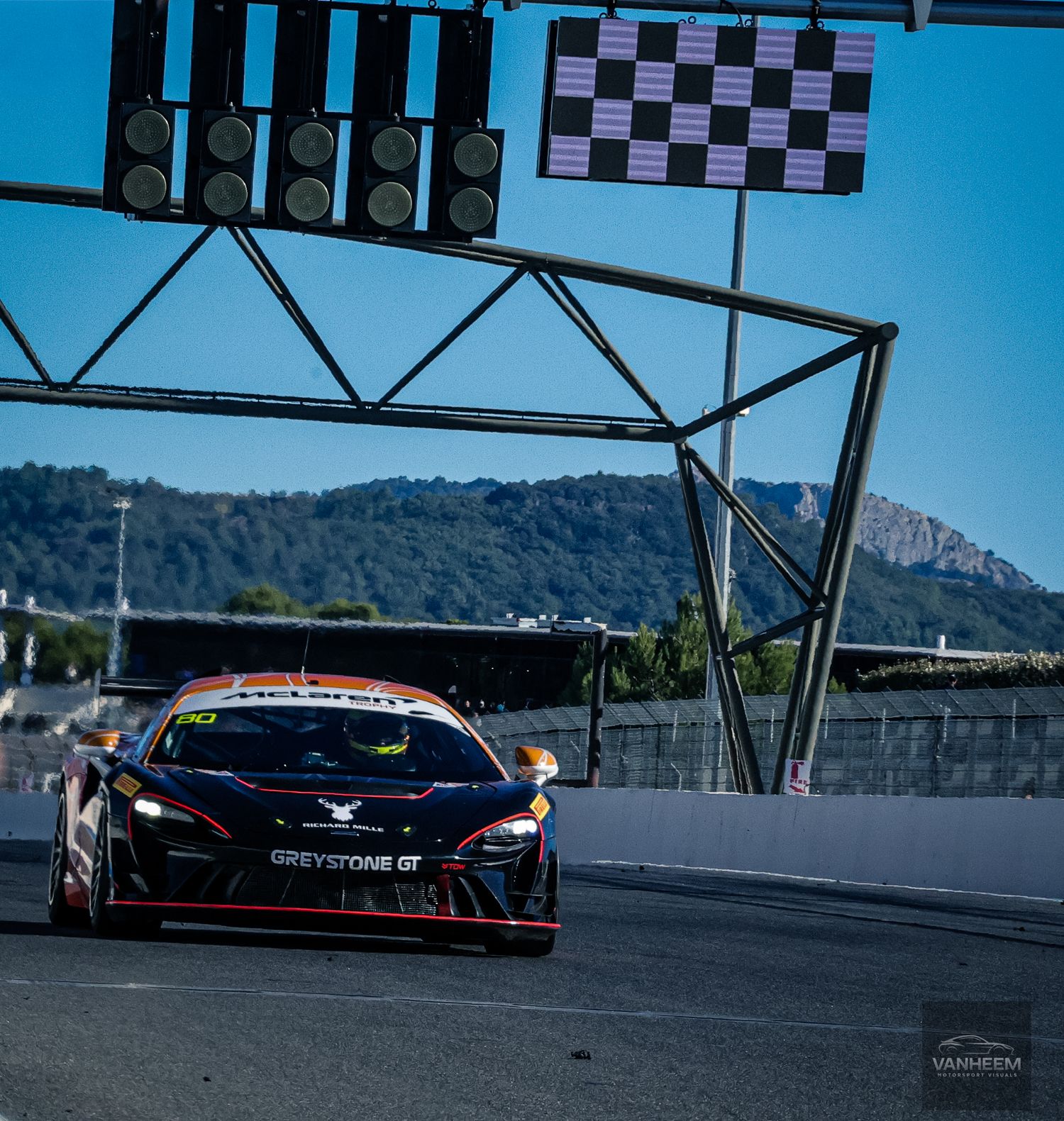 Black racing car speeds past checkered flag on racetrack with mountain landscape in background under blue sky.