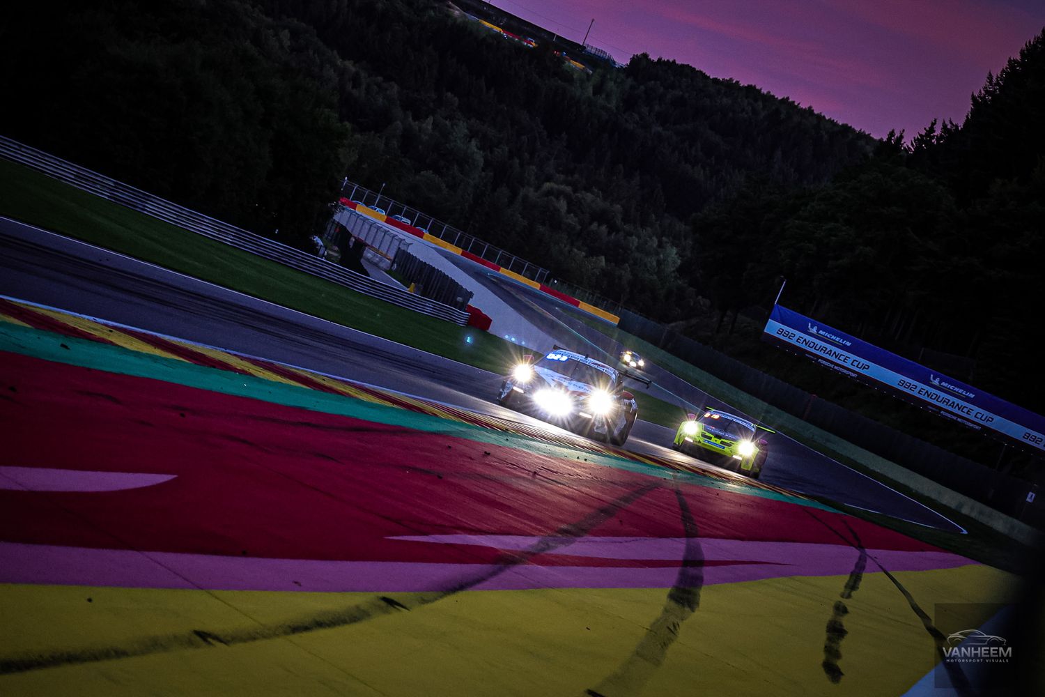 Racing cars speed through a curved track at dusk with colorful track markings and purple sky overhead.