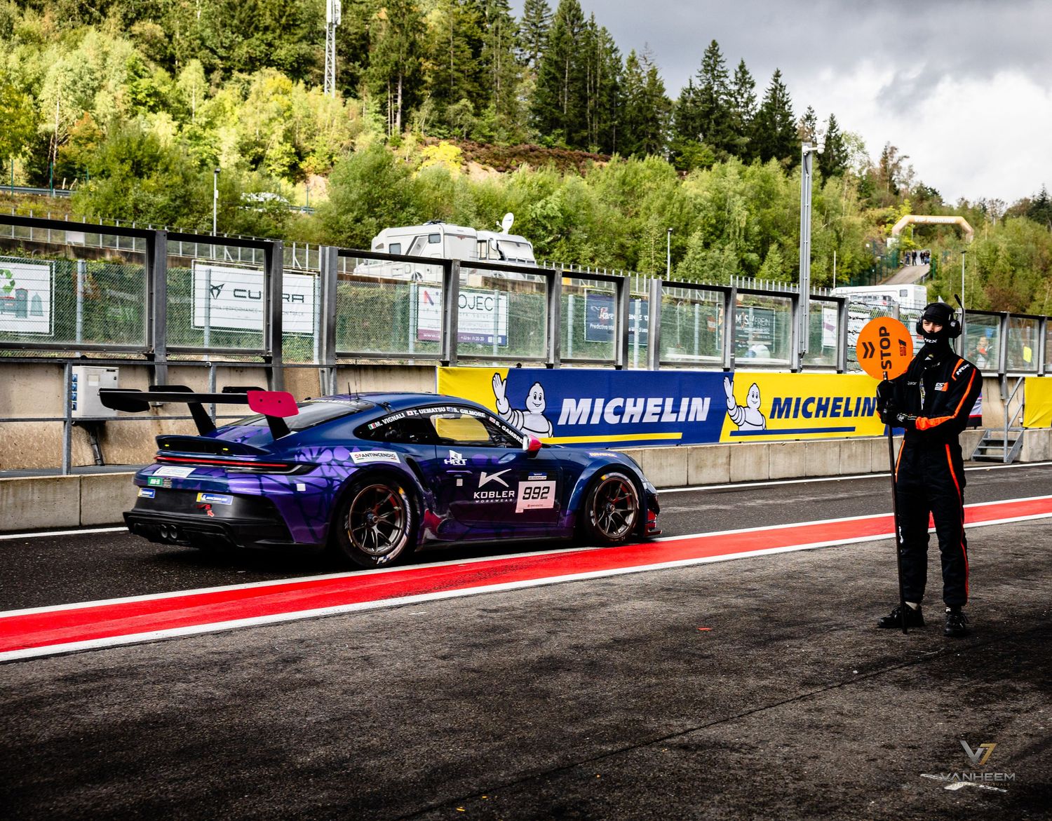 Purple Porsche GT3 race car on Spa-Francorchamps circuit pit lane with Michelin signage and marshal in background.