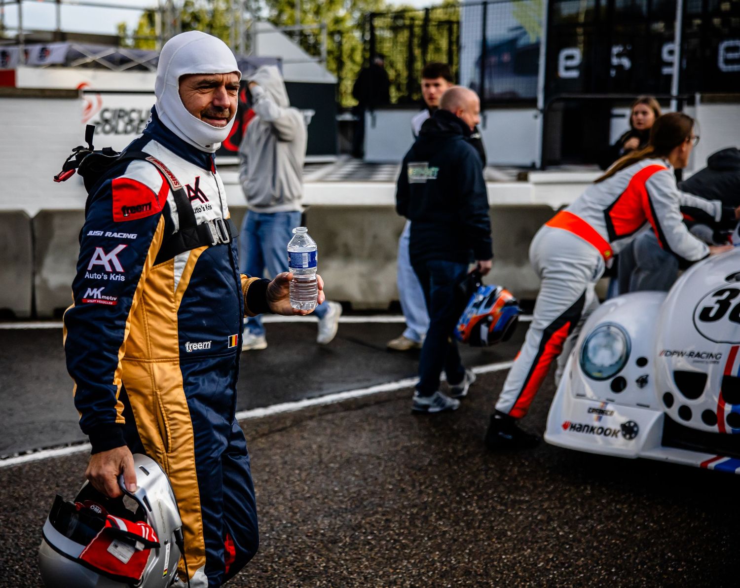 A racing driver in a gold and black firesuit holds a helmet at a karting event with team members and karts in the background.