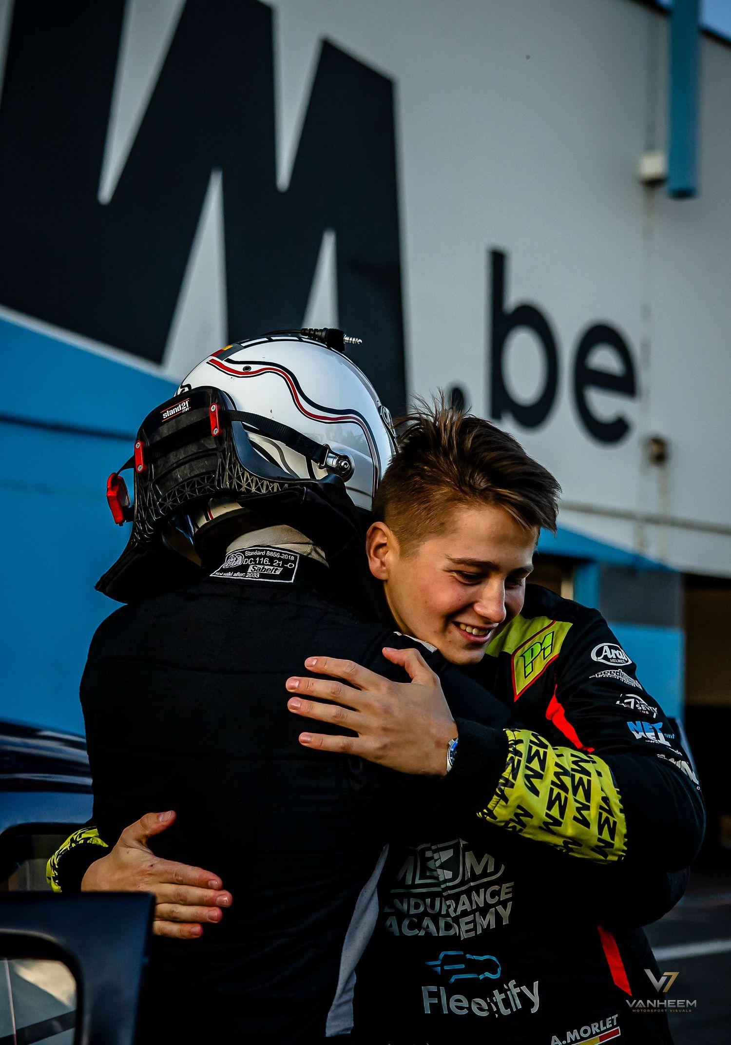 Young racing driver in M Endurance Academy gear shares a celebratory hug with a helmeted teammate at a motorsport event.