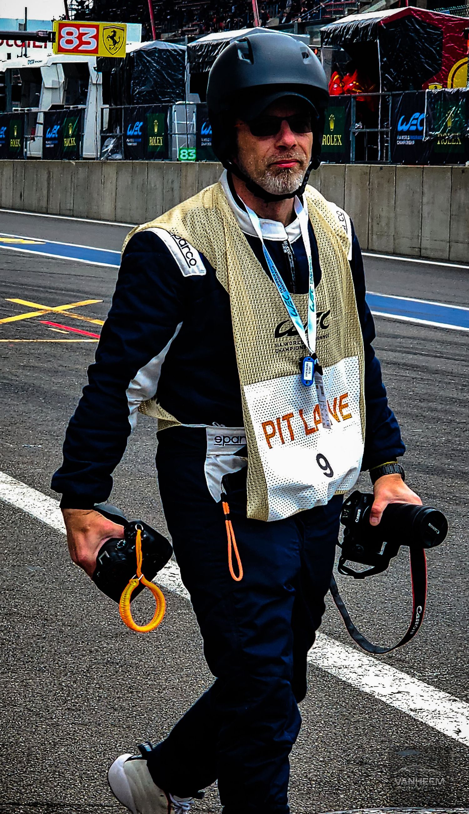 A photographer wearing a pit lane vest and helmet walks through a racing circuit pit lane carrying two cameras.