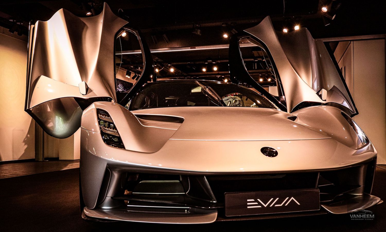 Lotus Evija electric hypercar with dramatic dihedral doors open, showcased under dramatic lighting at an auto exhibition.