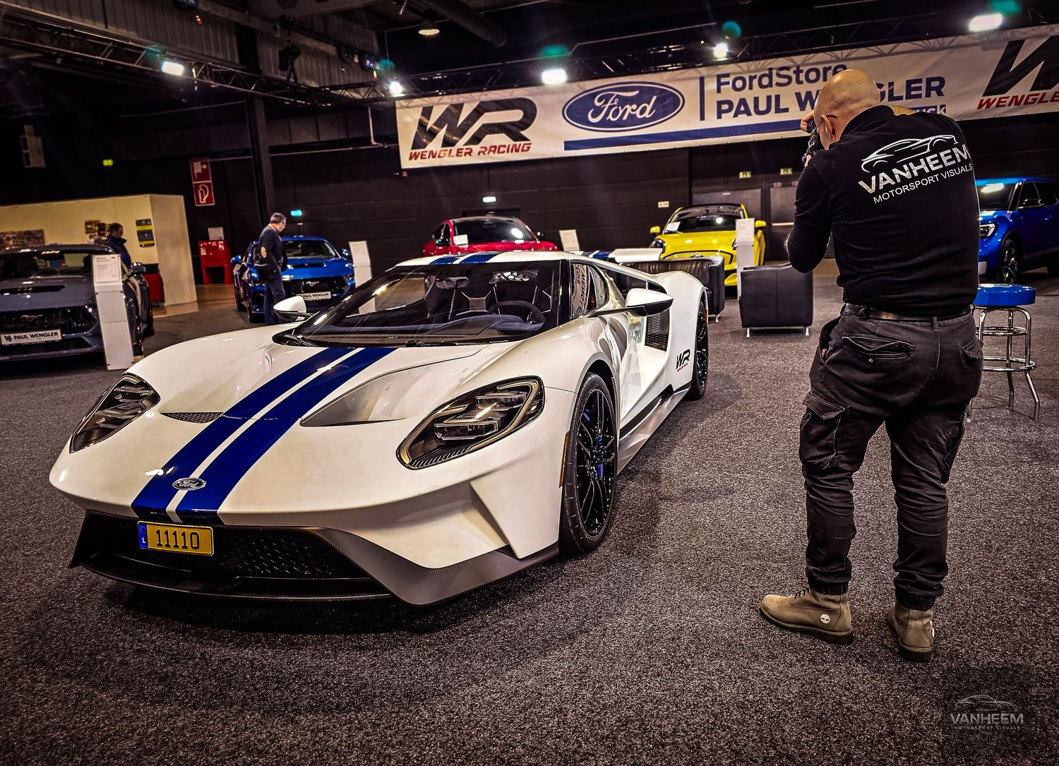 White Ford GT race car with blue stripes on display at an indoor auto show, with a man in black standing nearby.