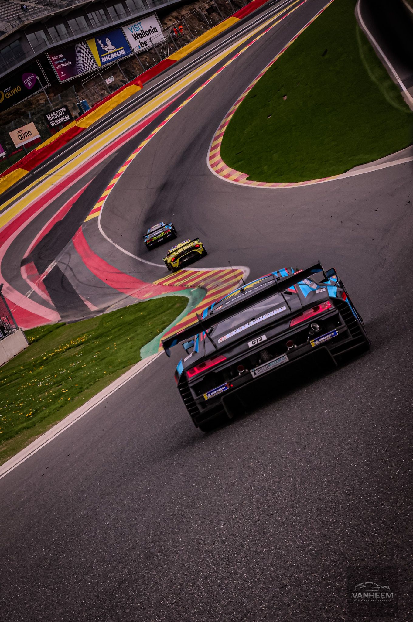 Racing cars navigate a winding circuit at Spa-Francorchamps, with a black GT car leading on the iconic track.