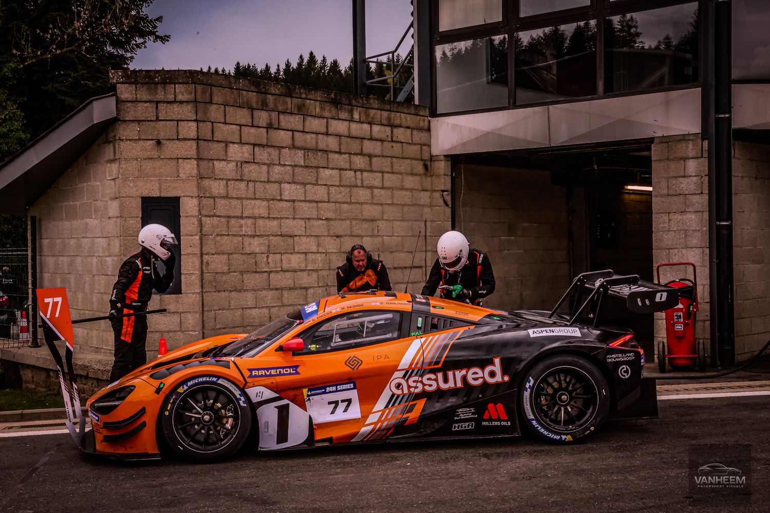 Orange and black #77 GT racing car in pit lane with crew members in orange suits at a motorsport circuit.