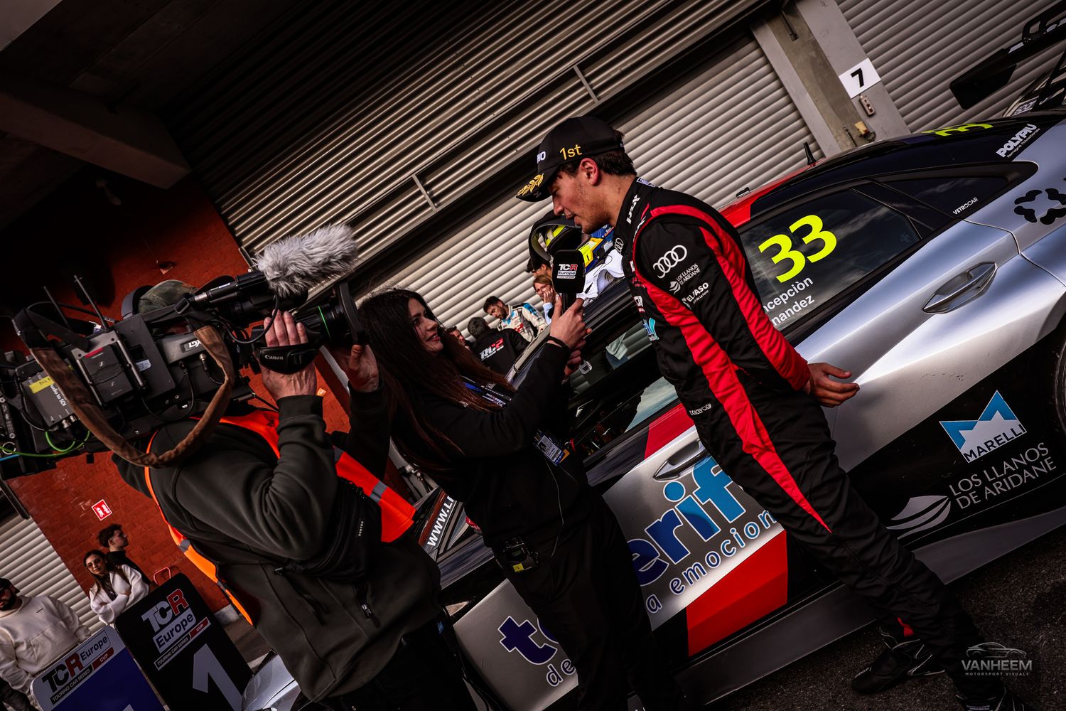 Racing driver in black suit being interviewed by camera crew in pit garage near car number 33.